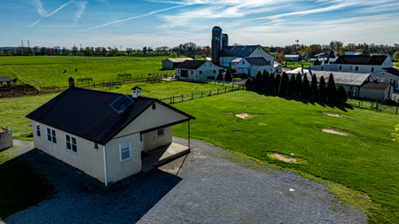 An Amish one room school house sits on a gravel area, surrounded by expansive green fields, barns, and distant structures under a bright blue sky.の写真素材