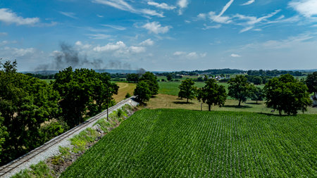 A steam locomotive chugs along tracks surrounded by lush green fields and trees on a bright day. The landscape features a clear blue sky with wispy clouds, enhancing the scenic view.の写真素材