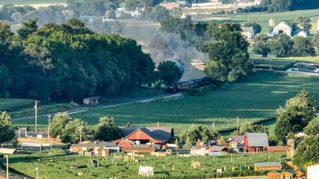 A vintage train chugs along a scenic route, billowing smoke as it passes through lush green fields and groves. Nearby, small farmhouses and barns dot the landscape under a clear sky.の写真素材
