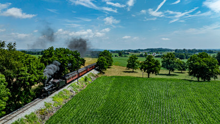 A steam locomotive chugs along tracks surrounded by lush green fields and trees on a bright day. The landscape features a clear blue sky with wispy clouds, enhancing the scenic view.の写真素材