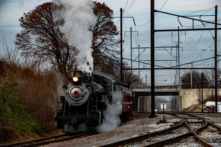 A vintage steam locomotive emits white smoke while pulling a passenger car on railway tracks. The...