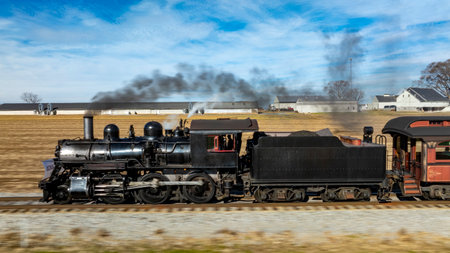 A vintage steam locomotive powers along the tracks, sending plumes of smoke into the blue sky. Surrounding fields and farmhouses enhance the picturesque countryside setting.の写真素材