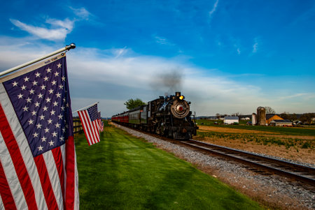 A vintage steam train chugs along scenic tracks bordered by green fields and American flags. The atmosphere is bright and inviting during a clear afternoon with a blue sky.の写真素材