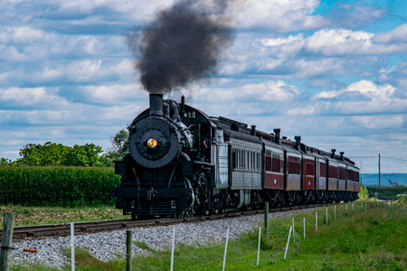 A vintage steam train puffs black smoke as it moves along the railway, surrounded by lush green cornfields and under a bright blue sky with fluffy clouds, creating a picturesque countryside view.の写真素材