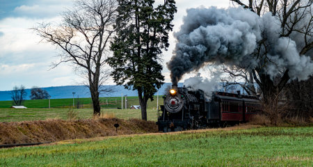 A vintage steam train chugs along railway tracks in a vast rural area, emitting thick plumes of smoke against a backdrop of dark clouds. Nearby fields exhibit golden crops.の写真素材
