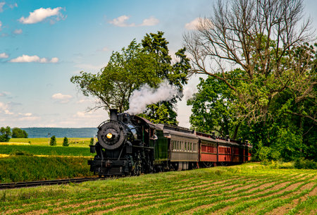 A steam train chugs along railway tracks bordered by vibrant green fields. The sun shines down, illuminating the historic engine and colorful carriages amid a tranquil landscape.の写真素材