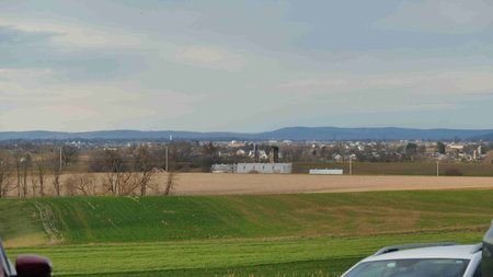 Wide view of a tranquil rural landscape reveals expansive green and brown fields. In the distance, farm buildings and a small town are visible, framed by gentle hills.の写真素材