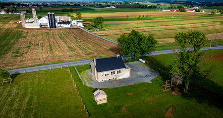 An Amish one room school house sits on a gravel area, surrounded by expansive green fields, barns, and distant structures under a bright blue sky.の写真素材