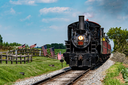 A classic steam train moves along the tracks in lush green countryside. American flags line the pathway, adding color to the bright blue sky and warm sunlight.の写真素材