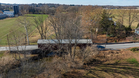 A covered bridge stands along a minor road, surrounded by bare trees and grassy fields. The serene...