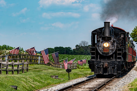 A classic steam train moves along the tracks in lush green countryside. American flags line the...