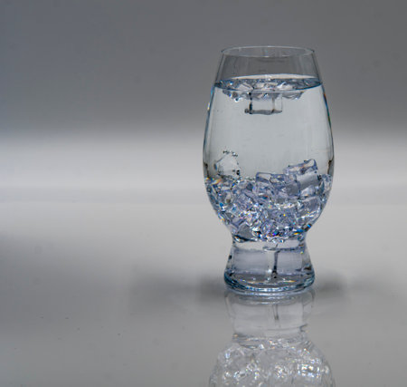 A clear glass filled with water and ice cubes sits on a smooth, reflective surface. The light highlights the clarity of the liquid and the ice, creating a refreshing appearance.の写真素材