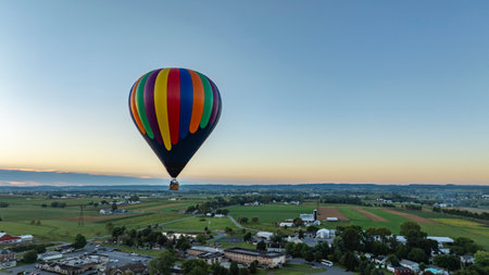 A vibrant hot air balloon ascends into the early morning sky, casting a shadow over lush farmlands and rolling hills. The tranquil scene captures the beauty of nature at dawn.の写真素材