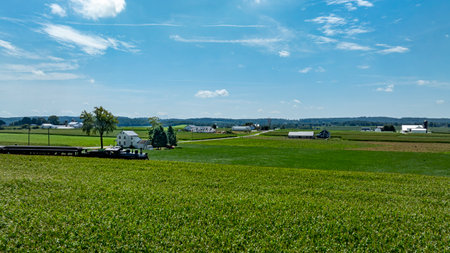 Expansive green fields stretch across the landscape, dotted with farmhouses. Rolling hills and a clear blue sky complement this serene rural setting on a sunny day.の写真素材