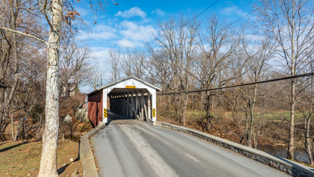 A charming covered bridge spans a tranquil road in a rural setting. The sunny winter day highlights the bare trees, making this scene picturesque and serene.の写真素材