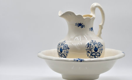 A decorative ceramic pitcher is placed beside a matching basin, both adorned with intricate blue floral designs. The items are positioned on a smooth white surface, highlighting their craftsmanship.の写真素材