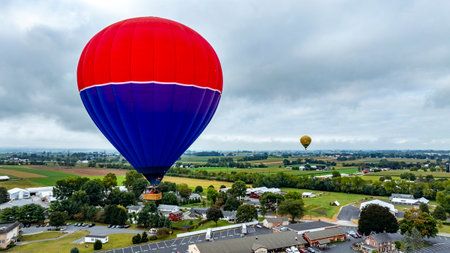A vibrant hot air balloon floats high above open fields and quaint houses, with another balloon visible in the distance, creating a stunning view of a rural landscape.の写真素材