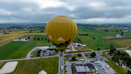 A vibrant hot air balloon floats gracefully over a picturesque rural landscape filled with green fields and scattered buildings. The cloudy sky adds a serene atmosphere to the scene.の写真素材