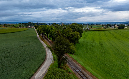 A winding road separates lush green fields while leading towards a small rural village. The sky is filled with clouds, hinting at an approaching storm.の写真素材