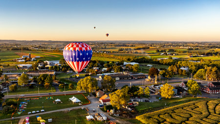 Hot air balloons soar above green fields and quaint buildings in a small town during the early morning hours. The vibrant colors stand out against the serene landscape.の写真素材