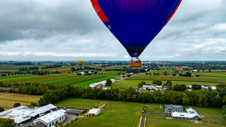 A vibrant hot air balloon ascends above lush green fields and quaint buildings, capturing the beauty of the morning in a serene countryside setting.の写真素材