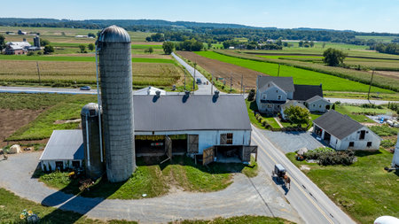 A large Amish barn with silos is situated near a winding road, surrounded by lush green fields and a...