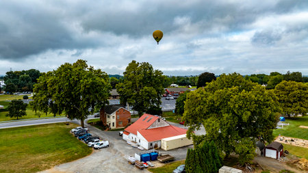 A hot air balloon ascends gracefully above a rural area, surrounded by lush trees and a charming building. The overcast sky adds a unique ambiance to the scene.の写真素材