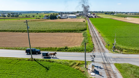 An Amish horse-drawn carriage moves down a rural road, with a vehicle close by, while a plume of smoke rises in the background near farm buildings on a sunny day.の写真素材