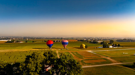In the early morning light, vibrant hot air balloons stand ready for ascent amidst expansive fields and an idyllic countryside view. The calm atmosphere sets the stage for an adventurous flight.の写真素材