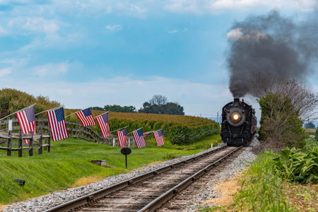 A classic steam train makes its way through a picturesque rural landscape, flanked by American flags. The scene captures the charm of vintage rail travel on a clear day.の写真素材