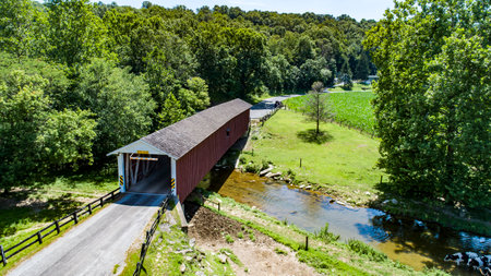 A quaint covered bridge spans a gentle stream, nestled among vibrant trees and fields.の写真素材