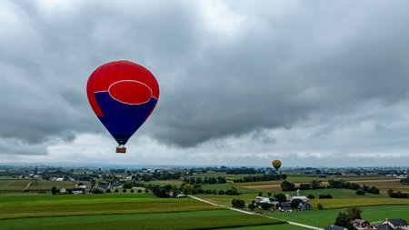 A vibrant hot air balloon floats high above open fields and quaint houses, with another balloon visible in the distance, creating a stunning view of a rural landscape.の写真素材