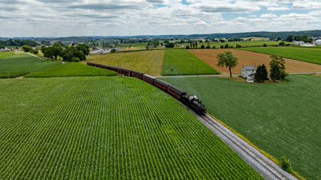 A steam train winds its way through vibrant green fields and rolling hills in Pennsylvania, with a quaint farmhouse visible nearby under a cloudy sky, showcasing rural charm.の写真素材