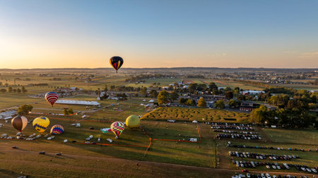 Colorful hot air balloons ascend into the evening sky over a festival ground surrounded by green fields and parking areas, capturing the essence of a lively rural celebration.の写真素材