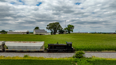 A black locomotive moves along the tracks beside lush green fields. It passes a quaint farmhouse, with clouds floating overhead on a summer afternoon, showcasing rural life.の写真素材