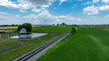A vintage train runs alongside a lush green field and a calm pond, under a bright blue sky with fluffy clouds. The rural scenery showcases the beauty of the countryside.の写真素材