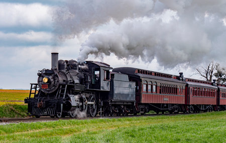 A vintage steam train chugs along railway tracks surrounded by lush green fields under a clear blue sky. The steam billows into the air, creating a picturesque moment.の写真素材