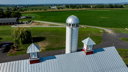 A large farm features a striking silver silo amidst expansive green fields under a clear sky. The late afternoon sunlight enhances the serene landscape, creating a peaceful rural atmosphere.の写真素材