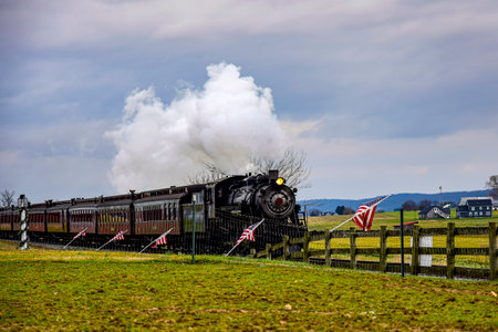 Historic steam locomotive pulls vintage passenger cars along tracks while puffs of steam rise against a cloudy sky. The setting features a green field and wooden fence nearby.の写真素材