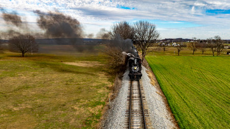 A vintage steam locomotive chugs along the railway track, emitting smoke as it travels through a serene rural landscape with green fields and bare trees under a blue sky.の写真素材