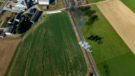 A plume of smoke emerges from the railway tracks surrounded by lush green fields and farm buildings under a bright blue sky. The scene captures rural life in motion.の写真素材