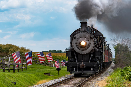 A classic steam train makes its way through a picturesque rural landscape, flanked by American flags. The scene captures the charm of vintage rail travel on a clear day.の写真素材
