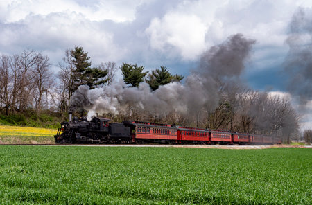 A vintage steam locomotive pulls a red passenger car along a scenic route. The train emits plumes of smoke against a backdrop of green fields and overcast skies, capturing a moment of nostalgia.の写真素材