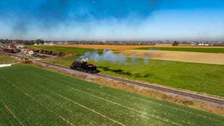 A steam train puffs along a rural track, releasing clouds of smoke as it moves through picturesque farmland. Green fields and a blue sky create a serene landscape.の写真素材