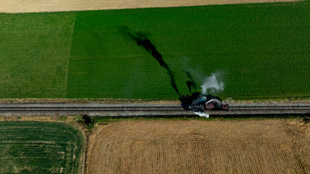 A vintage steam train moves along railway tracks, producing plumes of smoke as it crosses vibrant green and golden fields under clear blue skies. The trains journey showcases rural landscapes.の写真素材