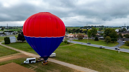 A vibrant hot air balloon is being set up for a flight in a scenic rural area. Surrounding fields and small towns create a picturesque backdrop on this overcast day.の写真素材