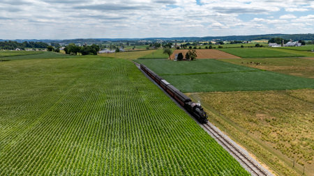 A vintage steam train moves along railway tracks surrounded by vibrant green fields and farmland. The open landscape features rolling hills and a clear blue sky with scattered clouds.の写真素材