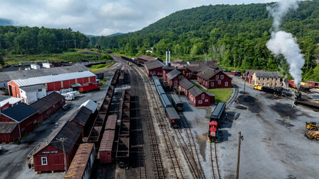 A well-maintained railway yard features vintage trains parked alongside rustic red buildings. Lush green hills surround the area, with steam billowing in the background.の写真素材
