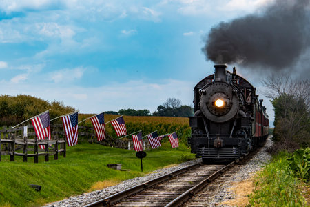 A classic steam train makes its way through a picturesque rural landscape, flanked by American flags. The scene captures the charm of vintage rail travel on a clear day.の写真素材