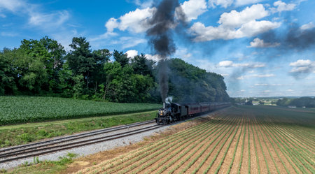 A Black Steam Locomotive Billowing Thick Smoke Pulls Red Passenger Cars Through Lush Green Fields And Forested Areas, Under A Clear Blue Sky With Puffy Clouds, Creating A Picturesque Countryside Journey.の写真素材